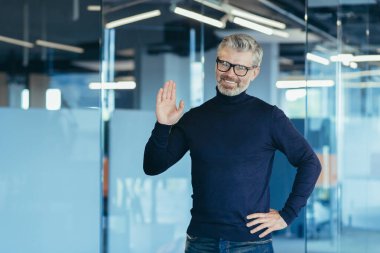 Portrait of successful financier in modern office, man looking at camera and smiling holding hand up greeting gesture, gray haired senior experienced businessman with beard and glasses, investor owner