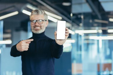 Senior successful gray-haired businessman in office, showing phone screen to camera with white screen background, man pointing with finger on copy space