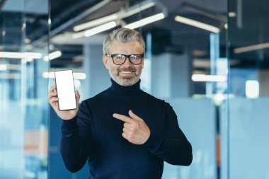 Senior successful gray-haired businessman in office, showing phone screen to camera with white screen background, man pointing with finger on copy space
