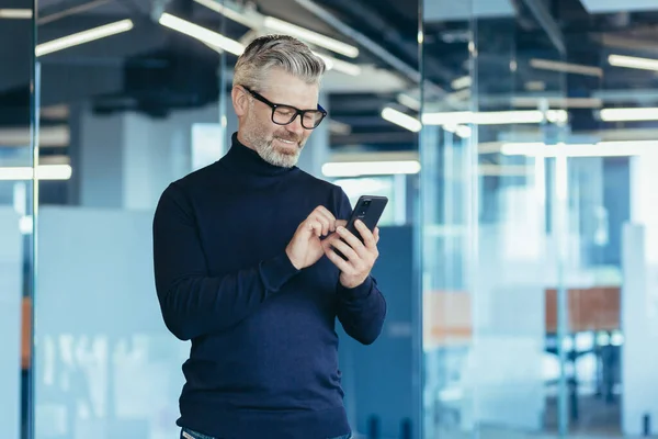 Senior gray-haired businessman in office using phone, happy investor reading message and smiling, broker at work looking at window in glasses