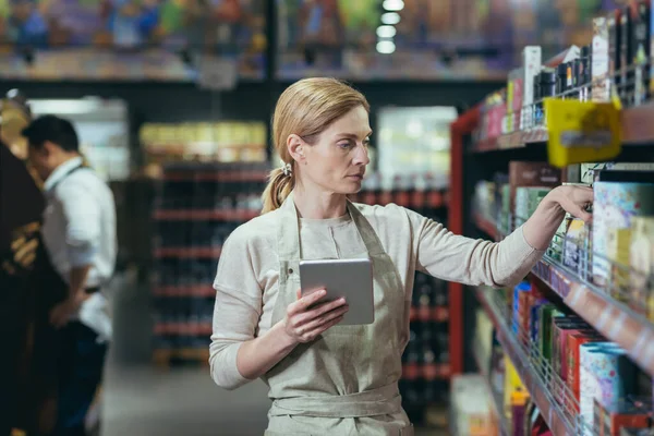 A woman seller in a supermarket uses a tablet computer to count the remaining goods, colleagues conduct an inventory in the grocery department among the shelves with goods