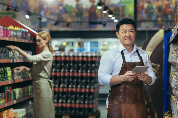 Portrait of a successful and happy Asian seller in a supermarket, a man in an apron is looking at the camera and smiling, the store manager is holding a tablet computer for product review