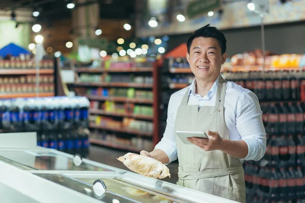 Portrait of Asian store manager, man with tablet checking expiration date of products, seller smiling and looking at camera near refrigerator in supermarket