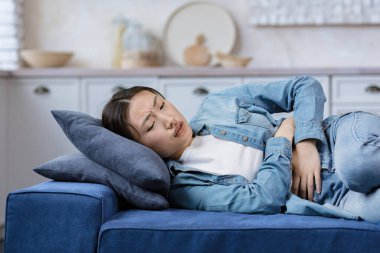 Young beautiful woman sick alone at home lying on bed, Asian girl holding hands on stomach, having severe stomach pain lying on sofa in living room