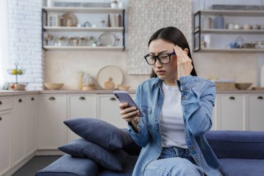 Young beautiful asian upset woman sitting at home in living room on sofa wearing glasses, reading message on mobile phone frustrated, sad in depression