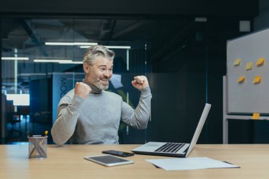 Successful and happy owner senior gray-haired investor mature businessman working in modern office using laptop, man celebrating victory triumph, holding hands up and smiling, success gesture