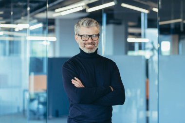 Portrait of successful businessman in modern office with crossed arms, gray haired male investor smiling and looking at camera, boss in glasses and jeans