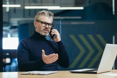 Senior and mature boss businessman working inside office building, man thoughtful and serious talking on the phone, gray haired investor in glasses working using laptop