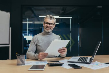 Portrait of mature experienced financier, man working with documents inside office building, businessman smiling and looking at camera holding documents and bills, boss working using laptop