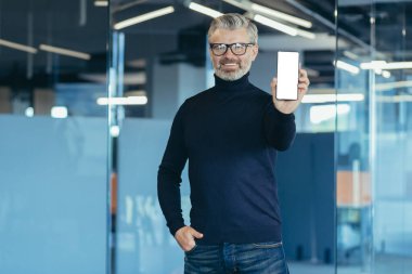 Senior experienced gray haired businessman working inside office, portrait of man with phone, mature investor smiling and looking at camera, showing white mobile phone screen