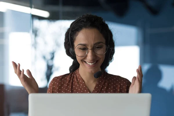 Close-up photo of young beautiful curly Arab woman talking on video call, looking at laptop web camera and smiling using headset for communication, successful businesswoman working in office