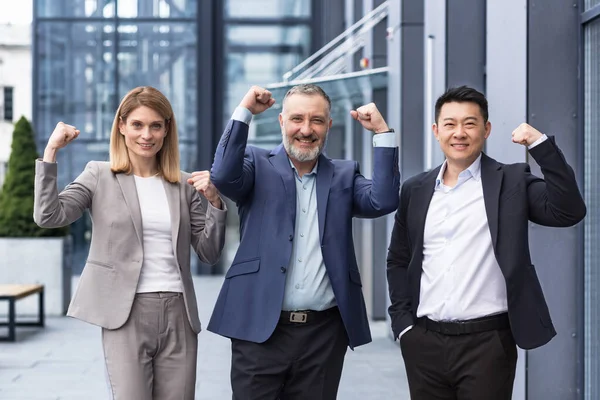 Successful diverse business team, man and woman outside office building smiling and looking at camera, portrait of business people in business clothes, colleagues holding hands up and rejoicing