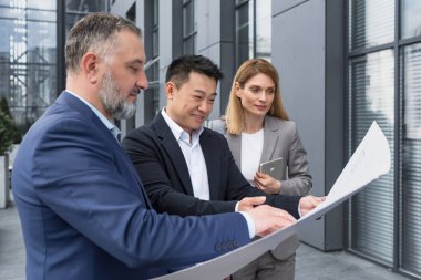 Diverse group of business people outside office building, discussing and talking man and woman discussing new construction plan, team of business people holding and looking at construction project