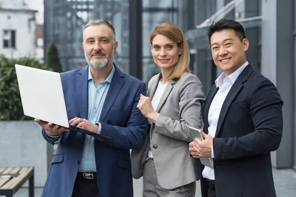 Portrait of three diverse business colleagues, business group outside office building, asian man and woman looking at camera and smiling, dream team with laptop
