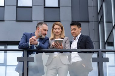 Three colleagues, diverse business group, business people outside office building talking and discussing work process, looking at tablet computer thoughtfully