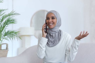 Young beautiful Muslim woman in hijab at home talking on the phone, woman in living room smiling and happy holding smartphone in hand chatting with friends