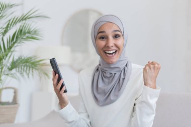 Portrait of young beautiful woman in hijab, arab muslim woman looking at camera and smiling sitting on sofa in living room, holding phone in hands happy success victory and good news