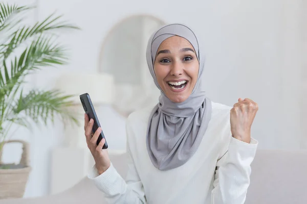 Portrait of young beautiful woman in hijab, arab muslim woman looking at camera and smiling sitting on sofa in living room, holding phone in hands happy success victory and good news