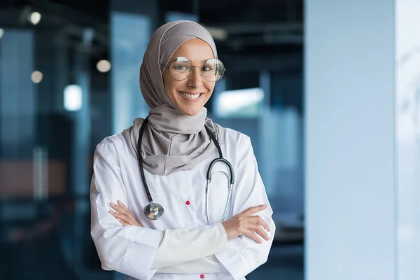 Portrait of Arabic female doctor in modern clinic, Muslim woman wearing hijab glasses and white medical coat working in hospital office, smiling and looking at camera with crossed arms