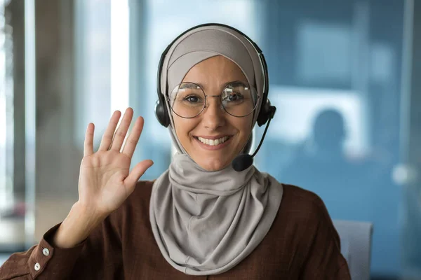 Close-up photo portrait of young beautiful Muslim business woman, tech support worker in hijab and glasses, customer service employee smiling and looking at camera, using headset , video call.