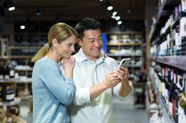 Young diverse couple man and woman in liquor department choosing wine, smiling and happy using smartphone to identify wine, using app to scan products