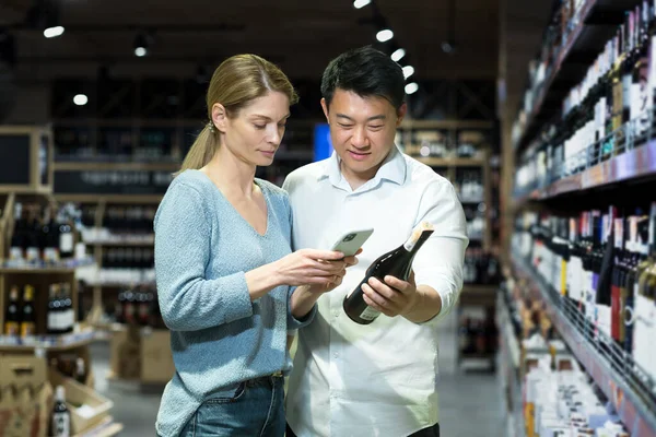 Diverse young married couple man and woman shoppers in supermarket, choosing alcohol wine, using app on phone to identify and scan wine bottle products