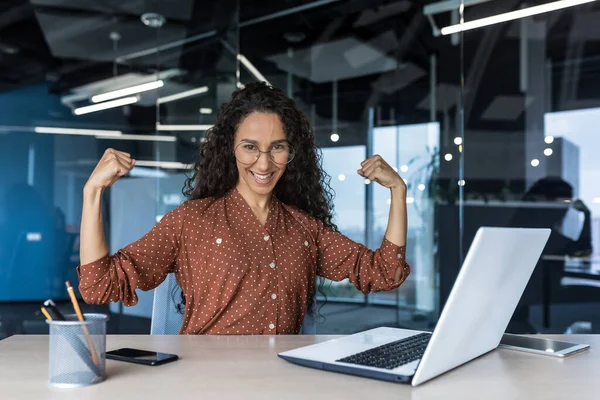 Portrait of a strong business woman female worker working inside office building, sitting a desk and using a laptop, smiling and looking at the camera holding hands up gesture superpower and success