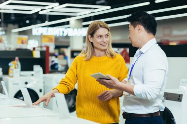 Asian consultant salesman in electronics and household appliances store, selling a working machine to a woman, recommending and approving the choice