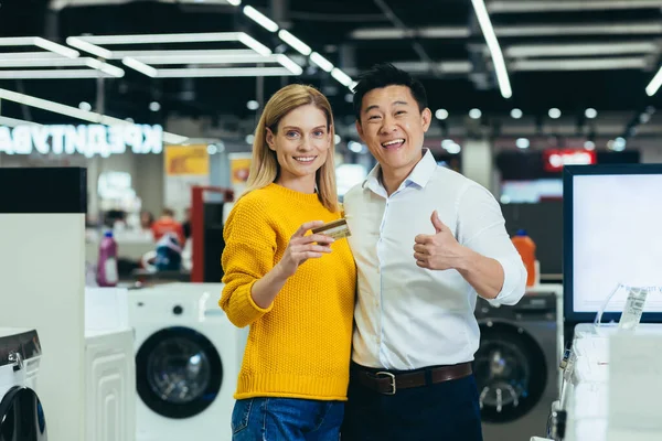 Portrait of diverse family couple Asian man and woman shopping in supermarket, electronics and household appliances, looking at camera and smiling, satisfied with bank credit card