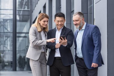 Diverse business group, three male and female workers outside office building cheerfully smiling and happy watching video on phone