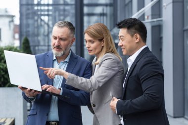 Successful business team, three colleagues businessman and businesswoman outside office building discussing current plans and management, looking at laptop screen, discussing and consulting