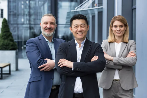 Successful diverse business team, three workers smiling and looking at camera, dream team with asian boss outside office building, colleagues in business suits