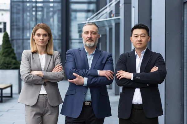 Successful and serious diverse team of three business people, man and woman focused looking at camera with arms crossed, portrait of co-workers outside office building