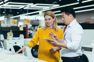 Asian consultant salesman in electronics and household appliances store, selling a working machine to a woman, recommending and approving the choice