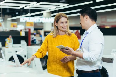 Asian consultant salesman in electronics and household appliances store, selling a working machine to a woman, recommending and approving the choice