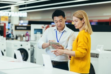 Asian consultant salesman in electronics and household appliances store, selling a working machine to a woman, recommending and approving the choice
