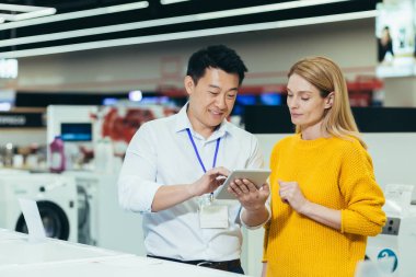 Asian consultant salesman in electronics and household appliances store, selling a working machine to a woman, recommending and approving the choice
