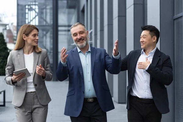 A diverse team of IT specialists, senior and experienced engineers managers team leaders, a group of three workers happily strolling outside an office building, colleagues in business suits.