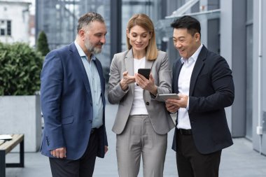 three IT specialists entrepreneurs outside office building watching video on colleagues phone, happy and smiling resting on break from work