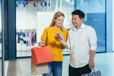 Diverse couple Asian man and blonde woman in clothes supermarket, looking at mobile phone screen, choosing shopping, holding colorful shopping bags and gifts.