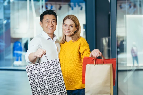 Young diverse family Asian man and blonde woman shopping in supermarket, portrait of couple shoppers looking at camera and smiling and holding colorful bags with goods and shopping.
