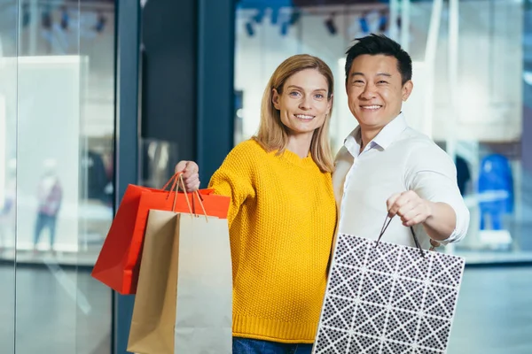 Young diverse family Asian man and blonde woman shopping in supermarket, portrait of couple shoppers looking at camera and smiling and holding colorful bags with goods and shopping.