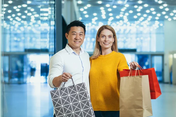 Young diverse family Asian man and blonde woman shopping in supermarket, portrait of couple shoppers looking at camera and smiling and holding colorful bags with goods and shopping.