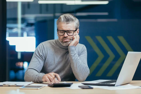 Upset and disappointed senior and mature financier, man working inside office building, calculating financial report with calculator, gray haired businessman investor in glasses thinking paperwork