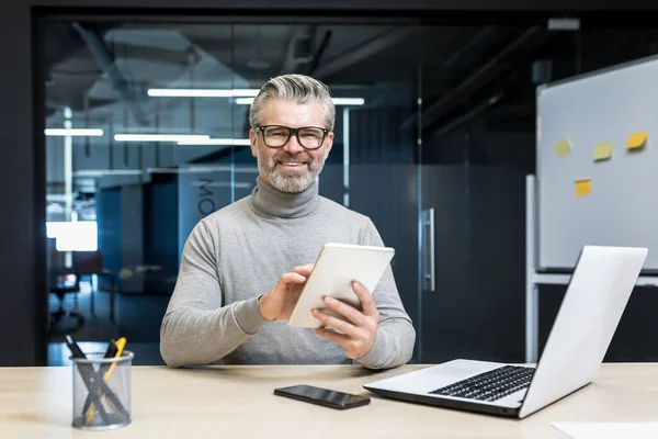 Portrait of successful programmer team leader, mature gray-haired engineer testing new app on tablet computer, businessman working inside modern office building, smiling and looking at camera