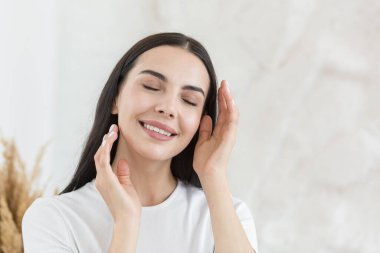 Cosmetology. Close-up portrait of young beautiful woman in white t-shirt brunette with clean white skin. She gently touches her face with her hands, closes her eyes, smiles.