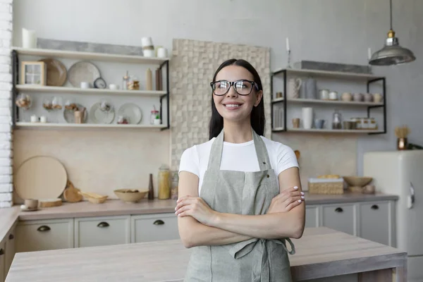 Portrait of a young beautiful woman cook, confectioner. She is standing in a beige apron and glasses in the kitchen, her arms crossed. He looks at the camera and smiles.