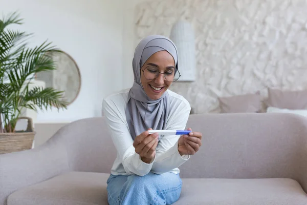 Happy and smiling Muslim woman in hijab holding pregnancy test at home, Arab woman happy with result sitting on sofa in living room.