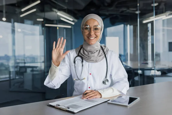 Video call online consultation, woman in hijab Muslim doctor looking at camera and waving greeting gesture, female doctor consulting patients remotely, working in modern clinic office