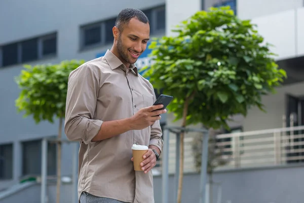 African American businessman smiling and happy walking outside the office building, holding a cup of hot drink during a break, and using the phone, browsing web pages and typing messages